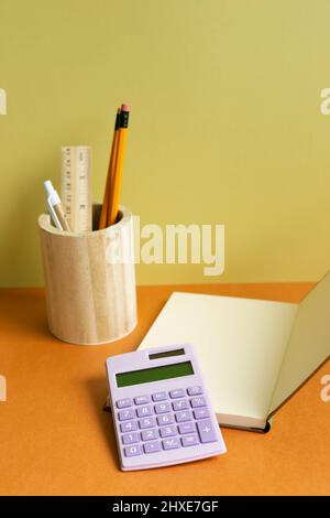 Notizbuch, Taschenrechner, Bleistifthalter auf orangefarbenem Schreibtisch. Gelber Wandhintergrund. Arbeitsbereich Stockfoto