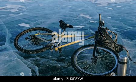 Mann und Fahrrad auf Eis. Er schaut auf das schöne Eis in den Rissen. Blick auf die erste Person auf dem Fahrrad. In der Nähe befindet sich eine Thermoskanne und ein Fahrradrucksack. Eis des Th Stockfoto