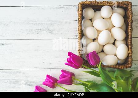 Weiße Eier in einem Korbkorb und ein Haufen burgunderfarbener Tulpen schließen sich auf hellem Holzhintergrund an, flach liegend, Kopierraum Stockfoto