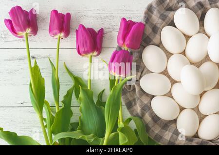 Weiße Eier und ein paar wunderschöne burgunderrote Tulpen aus der Nähe auf weißem Holzhintergrund, flach liegend Stockfoto