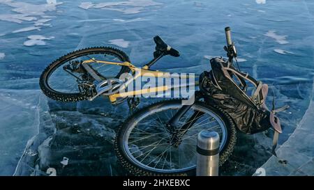 Mann und Fahrrad auf Eis. Er schaut auf das schöne Eis in den Rissen. Blick auf die erste Person auf dem Fahrrad. In der Nähe befindet sich eine Thermoskanne und ein Fahrradrucksack. Eis des Th Stockfoto