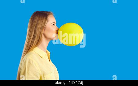 Seitenansicht eines glücklichen jungen Mädchens, das auf blauem Hintergrund einen gelben Ballon in die Luft sprengen kann Stockfoto