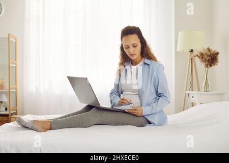 Schwangere, die mit einem Laptop auf dem Bett sitzt und sich im Notebook Notizen über die Schwangerschaft machen Stockfoto