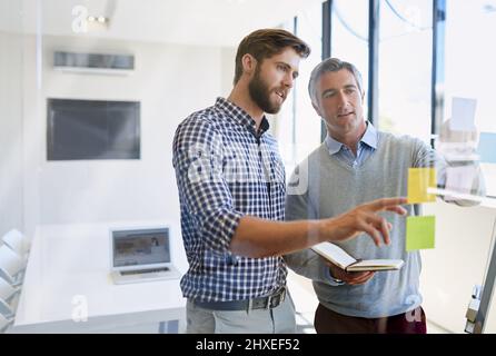 Sie glauben an ständige Innovationen, um ihre Kunden zu beeindrucken. Aufnahme von zwei männlichen Kollegen, die während eines Brainstormings Haftnotizen an einer Glaswand anordnen Stockfoto