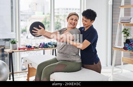 Haben heute eine positive und progressive Sitzung. Aufnahme einer jungen Physiotherapeutin, die ihrer erwachsenen Patientin mit einem Medizinball im Inneren beim Training hilft Stockfoto