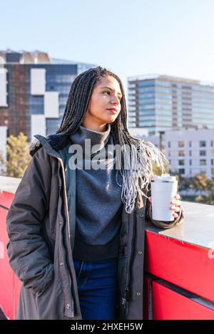 Verträumte hispanische Hündin mit Dreadlocks, die in der Nähe der Grenze mit weißen Thermoskannen mit heißem Kaffee in der Stadt mit Wohngebäuden stehen Stockfoto