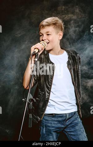 Porträt eines kaukasischen Teenagers in weißem T-Shirt, blauer Jeans und Lederjacke mit Mikrofon-Gesang auf dunklem Hintergrund. Konzept von Hooby und Glory Stockfoto