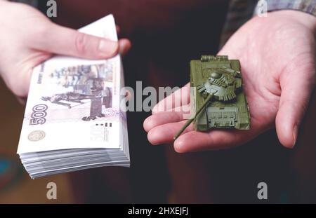 Russische Rubel Banknoten und Spielzeug Tank in den Händen während des Krieges in der Ukraine. Inflation, Sanktionen und der Sturz des russischen Wirtschaftskonzepts. Sinkende Bargeldeinsparungen und sinkendes Budget. Hochwertige Fotos Stockfoto