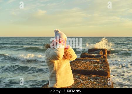 Eine Frau mit blondem Haar in einem Hut und einem Pelzmantel geht an einem Steinbrecher entlang des Meeres Stockfoto