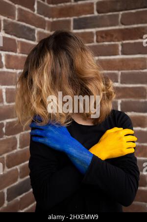 Unverkennbare Frau, mit Haaren bedecktes Gesicht, die ihre Hände erschöpft vor sich hält, gemalt in den Farben der ukrainischen Flagge. Krieg in der Ukraine. Su Stockfoto