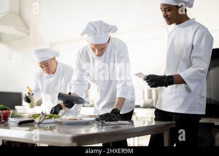 Mehrere Rassen Gruppe von Köchen, die Hauptgerichte beenden, während sie zusammen in der Küche arbeiten. Das Team bereitet Mahlzeiten für das Restaurant zu Stockfoto