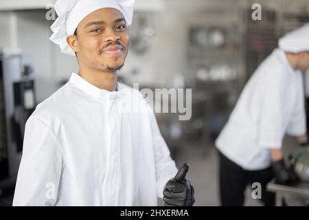 Porträt eines jungen lächelnden lateinamerikanischen Küchenchefs in Uniform in der professionellen Küche. Schöner Mann, der als Koch im Restaurant arbeitet Stockfoto