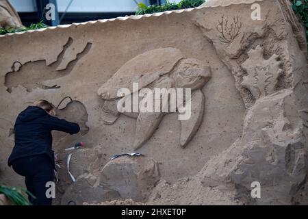 Binz, Deutschland. 11. März 2022. In der Sandskulptur-Show „What a wonderful world“ arbeitet eine Frau mit einer Schildkröte an einer Sandskulptur. Die Skulpturen zeigen Landschaften, Menschen und Fabelwesen. Quelle: Stefan Sauer/dpa/Alamy Live News Stockfoto