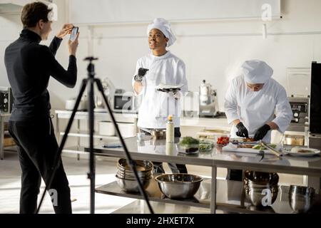 Mann zeichnet am Telefon den Prozess des Kochens von zwei Köchen in einer professionellen Küche auf. Lateinamerikanische und asiatische Köche bereiten Speisen für soziale Medien zu. Konzept des kulinarischen Videobloggens Stockfoto