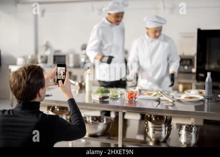 Mann zeichnet am Telefon den Prozess des Kochens von zwei Köchen in einer professionellen Küche auf. Lateinamerikanische und asiatische Köche bereiten Speisen für soziale Medien zu. Konzept des kulinarischen Videobloggens Stockfoto