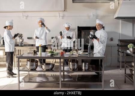 Gruppe von Köchen, die Telefone benutzen und während einer Pause in der Restaurantküche Kaffee trinken. Professionelles Personal ruht sich nach der Arbeit in der Küche aus. Ein Team von Köchen aus verschiedenen Rassen in Innenräumen Stockfoto
