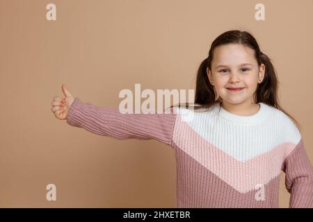 Porträt von jungen ziemlich glücklich lächelndes Mädchen mit langen dunklen Haaren in weiß, rosa Pullover stehen, zeigt Daumen nach oben Zeichen, Geste der Zustimmung auf braun Stockfoto