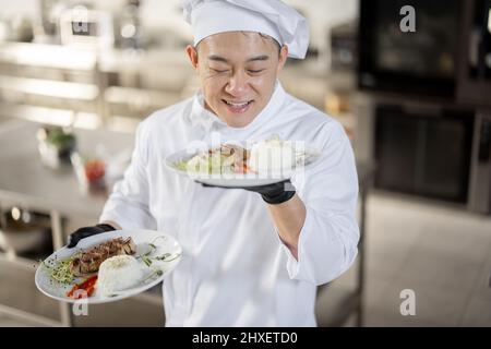 Porträt eines jungen glücklichen, gutaussehenden, lächelnden Küchenchefs in Uniform, der mit einer Fertigmahlzeit in der Küche steht. Glücklicher asiatischer Koch, der im Restaurant arbeitet. Konzept der beruflichen Tätigkeit und Haute Cuisine Stockfoto