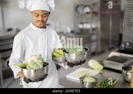 Portrait eines jungen Küchenchefs, der mit gesunden Zutaten steht und in der Restaurantküche kochbereit ist. Lateinischer Mann mit grünem Gemüse, der im Restaurant Mahlzeiten zubereitete Stockfoto
