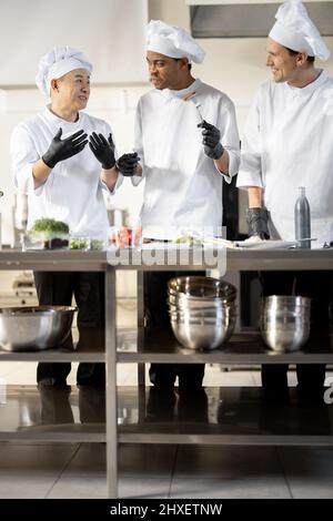 Drei Köche mit unterschiedlichen Nationalitäten verkosten gekochte Gerichte und erfinden neue Gerichte für ein Menü. Asiatischer Koch mit lateinischen und europäischen Köchen in Uniform, die zusammenarbeiten. Konzept der Teamarbeit und hohe Küche Stockfoto