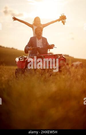 Junge fröhliche Jungvermählte haben Spaß, während genießt Hochzeit Fahrt mit Quad auf einem schönen Sonnenuntergang in der Natur. Reiten, Hochzeit, Natur, Aktivität Stockfoto