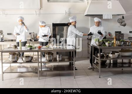 Gruppe von Köchen, die Telefone benutzen und während einer Pause in der Restaurantküche Kaffee trinken. Professionelles Personal ruht sich nach der Arbeit in der Küche aus. Ein Team von Köchen aus verschiedenen Rassen in Innenräumen Stockfoto