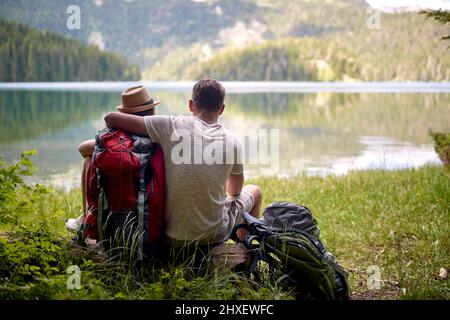 Paar auf Wanderung. Umarmen und sitzen vor dem See mit Rucksäcken. Wandern, Lifestyle, Freizeitkonzept Stockfoto