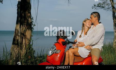 Motorroller Road Trip. Liebespaar auf rotem Motorrad in weißer Kleidung auf Waldweg. Gerade verheiratete Frau und Mann küssen, umarmen, sitzen auf dem Motorrad. Weddin Stockfoto