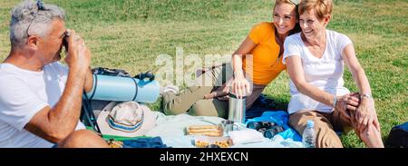 Älterer Vater, der ein Foto von seiner Familie machte, die auf einer Decke saß und ein Picknick machte Stockfoto