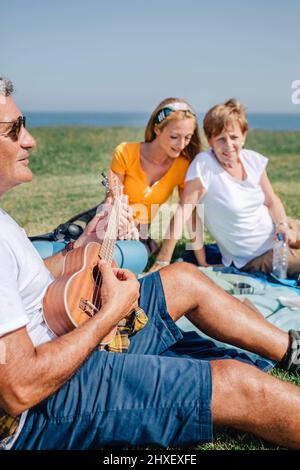 Älterer Mann spielt Ukulele für seine Familie und sitzt auf einer Decke während eines Ausflugs Stockfoto