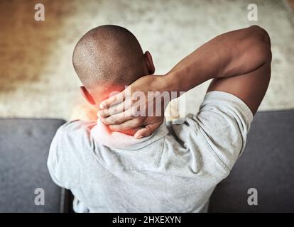 Ich muss letzte Nacht falsch geschlafen haben. Rückansicht eines nicht erkennbaren Mannes, der seinen Hals in Unannehmlichkeiten hielt, während er zu Hause auf einer Couch saß. Stockfoto