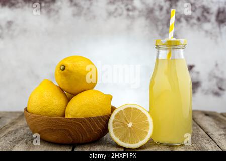 Flasche kalte Limonade mit Stroh und Holzschale mit Zitronen auf rustikalem Holztisch. Stockfoto