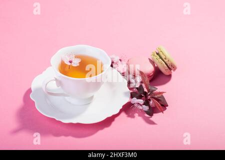 Tee mit rosa Frühlingskirschblüte in einer Porzellanbecher und einem Macaron serviert. Stockfoto