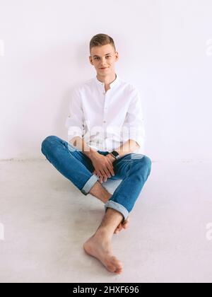 Hübscher Kerl in weißem Hemd und blauer Jeans sitzt auf einer Treppe drinnen. Mode, Stilkonzept Stockfoto