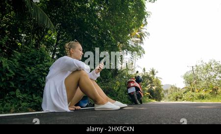 Frau auf rotem Motorrad in weißer Kleidung fahren auf Waldweg. Ein Mädchen kaukasischen Touristen sitzen, entspannen, ruhen, in der Nähe Roller. Verwenden Sie ein Smartphone, Stockfoto