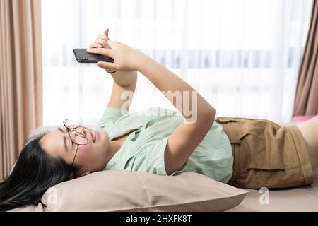Schöne Brille Asiatische Frau liegt auf dem Rücken, plaudert, tippt und schreibt mobil in ihrem Schlafzimmer mit einem pastellgrün-braunen Farbthema. Stockfoto
