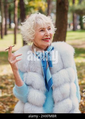 Lächelnd Senior elegant stilvolle modische Frau mit grauen Haaren in Pelzmantel im Freien Rauchen Zigarette. Ungesunder Lebensstil, Alter, positive Vibes Stockfoto