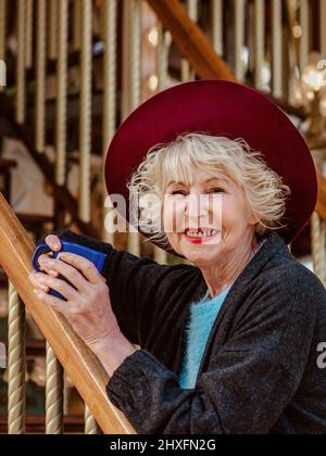 Ältere stilvolle Frau in dunkelgrauem Mantel, Hut und mit grauen Haaren, die am Karussell lächelnd stehen, Kaffee (Tee) trinken und das Leben genießen. Stockfoto
