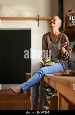 Shes lieben ihre neue Ernährung. Aufnahme einer attraktiven jungen Frau, die auf ihrer Küchentheke sitzt und beim Essen eines Salats lacht. Stockfoto