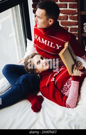 Paar in roten Pullovern, die sich im Bett entspannen und zusammen lesen. Stockfoto