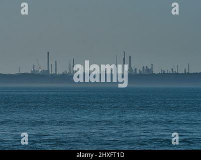 Eine zerklüftete Industrieanlage sticht durch den Dunst auf einer entfernten Landzunge über das strukturierte Wasser einer Bucht auf dem Solent in scharfes, sprödes Sonnenlicht Stockfoto