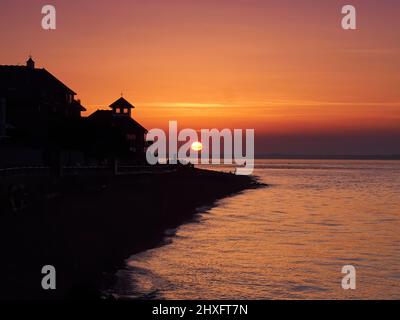 Ein farbenfroher Sonnenuntergang vom Cowes Beach aus, Sonnenlicht, das durch die Fenster eines Gebäudes am Meer kam und Menschen am Strand beschilderte. Stockfoto