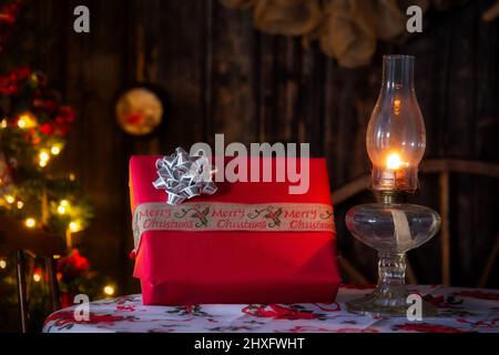 Ein Weihnachtsgeschenk in rotes Papier gewickelt und neben einer beleuchteten Öllampe im Retro-Stil auf einer Tischplatte verbeugt Stockfoto