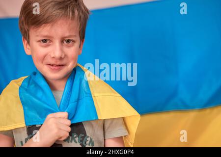 Junge hält ukrainische Flagge auf gelb-blauem Hintergrund in Händen. Die Kinder unterstützen die Ukraine mit Herzblut. Ukrainische Krise, russische Aggression in der Ukraine. Krieg Russlands gegen die Ukraine. Stockfoto