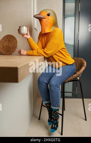 Eine Frau in einer Vogelmaske trinkt in der Küche eine Tasse Kaffee Stockfoto