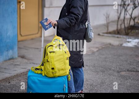 Evakuierung von Zivilisten, trauriges Kind mit der Flagge der Ukraine. Flüchtlingsfamilie aus der Ukraine, die die Grenze überquert. Hand mit einem Pass über dem Gepäck mit gelb-blauer Flagge. Beendet den Krieg, unterstützt die Ukraine Stockfoto