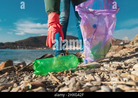 Ein Freiwilliger in Gummihandschuhen greift nach einer schmutzigen Plastikflasche, die am Meeresufer liegt. Nahaufnahme der Hände. Umweltverschmutzung. Stockfoto