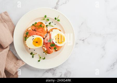 Sandwich mit köstlichen Belägen, geräuchertem Lachs, Eiern, Kräutern und Mikrogrüns Rettich, schwarzen Sesam über weißem Teller auf weißem Marmortisch Backgro Stockfoto