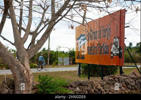 Mann, der in Giron in der Nähe von Havanna, Kuba, mit dem Fahrrad unterwegs ist; eine politische Plakatwand am Straßenrand in der Nähe von Havanna, Kuba. Stockfoto