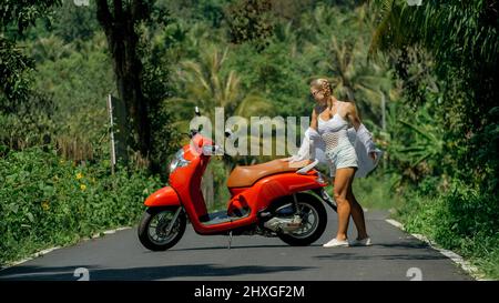 Frau auf roten Roller in weißen Kleidern fahren auf Waldweg. Tanzreise. Ein Mädchen kaukasischen Touristen in Sonnenbrillen tanzen, entspannen, Ruhe in der Nähe von Motor Stockfoto